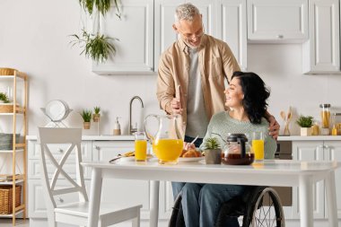 A man stands next to his wife in a wheelchair in their home kitchen. clipart