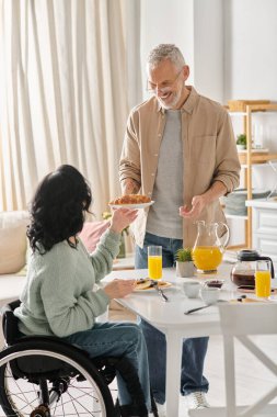 A man and a woman in wheelchair enjoying a meal together in their kitchen at home. clipart