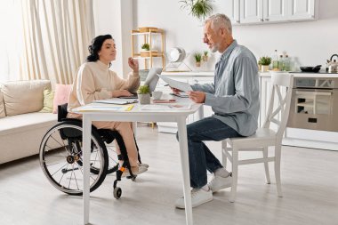 Bir adam evdeki mutfaklarında tekerlekli sandalyede oturan bir kadının yanında oturuyor..