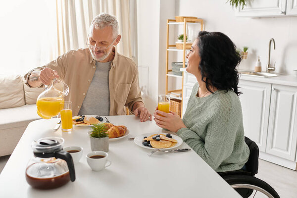 A disabled woman in a wheelchair and her husband enjoy a peaceful morning meal together at their kitchen table.