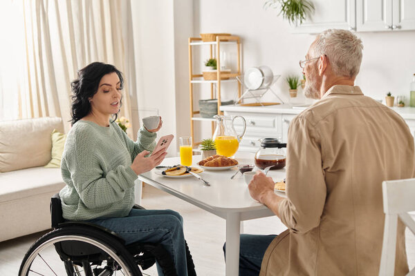 A man and woman in wheelchair share a moment at a kitchen table in their home, embracing to express their love and unity.