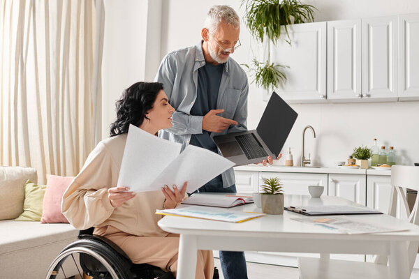 Женщина в инвалидной коляске сидит внимательно, используя ноутбук, погруженный в экран, в домашнюю кухню.