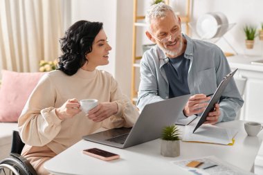 A man and woman, the woman in a wheelchair, sit at a table, engrossed in the screen of a laptop. clipart