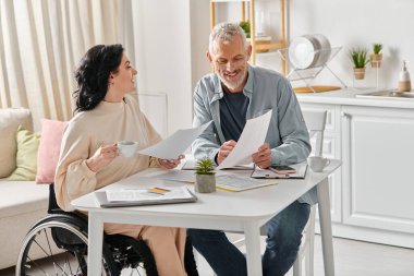 A woman in a wheelchair and her husband discussing budget at a table in their kitchen at home. clipart