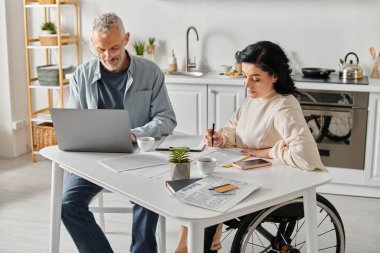 A husband and disabled wife sit at a kitchen table, working on a laptop together. clipart