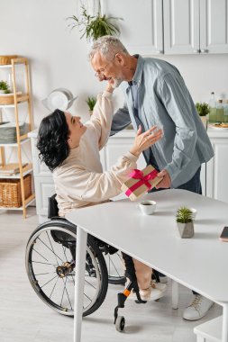 A disabled woman in a wheelchair touching her husbands hand lovingly in the kitchen at home, receiving present clipart