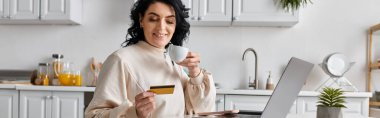 A happy woman holds a credit card while focused on her laptop in the kitchen. clipart