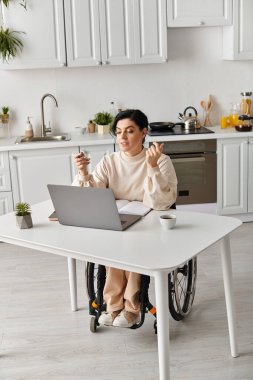 Tekerlekli sandalyedeki engelli bir kadın mutfaktaki bilgisayarında çalışıyor, masadaki dizüstü bilgisayara odaklanıyor..