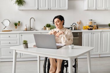 Tekerlekli sandalyedeki bir kadın mutfak masasında uzaktan çalışıyor, bağlı ve üretken kalmak için dizüstü bilgisayar kullanıyor..
