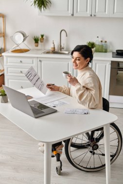 A woman in a wheelchair is sitting at a table in her kitchen, focused on working on her laptop remotely. clipart
