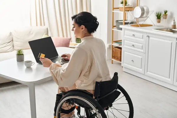 一位坐在轮椅上的残疾妇女正在厨房的笔记本电脑上远程工作 — 图库照片