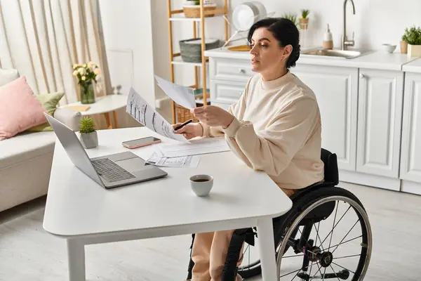 車椅子の障害のある女性が台所のテーブルでノートパソコンで作業しています — ストック写真