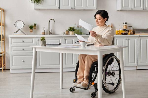 A woman in a wheelchair sits in her kitchen, deep in thought as she holds a piece of paper, focused on her creative work.
