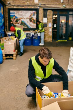 A man in safety gear sits by a cardboard box, pondering alongside young volunteers sorting trash.