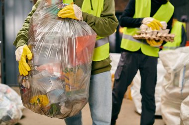 Two young volunteers in safety vests, gloves, and holding a huge bag of garbage while sorting trash.