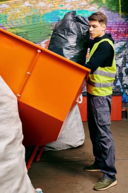 Young volunteer in gloves and safety vest stands next to dumpster, sorting trash in front of a wall.