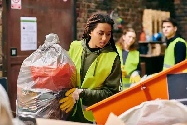 stock image A young woman in a green vest diligently collects trash in a plastic bag, joining other eco-conscious volunteers.