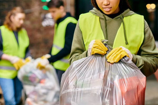 A young woman in gloves and safety vest holding a bag of garbage, recycling with fellow volunteers.