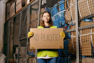 Young volunteer in gloves and safety vest holds a sign saying no plastic while sorting waste.