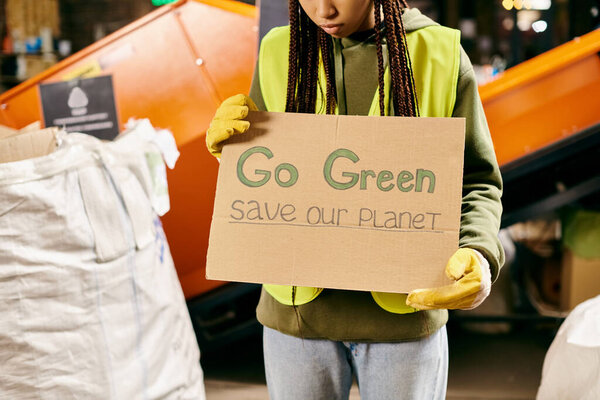 A young volunteer in gloves and a safety vest holds a sign urging to go green and save our planet.