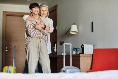 Senior lesbian couple share a tender hug in a luxurious hotel room. clipart