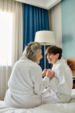 Two senior women, a loving lesbian couple, sit peacefully on top of a bed in a hotel room. clipart