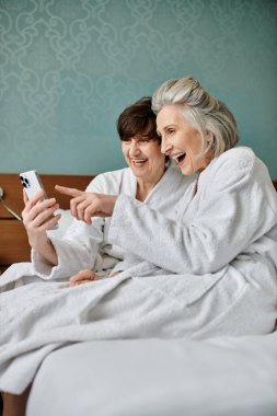 Yaşlı lezbiyen çift ve genç kadın cep telefonuyla yatakta eğleniyorlar..