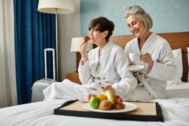 Senior lesbian couple serenely sitting on a cozy bed in a hotel room. clipart