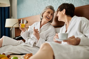 İki yaşlı kadın, samimi bir kucaklaşmada bir yatakta huzur içinde oturuyor..