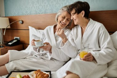 Two senior women sit closely on a bed, embodying love and tranquility. clipart