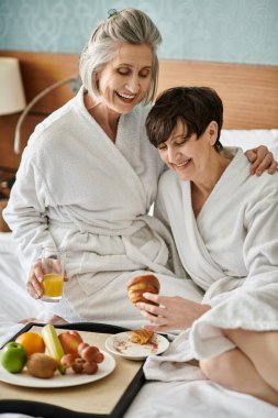 Senior lesbian couple shares a tender moment in a cozy bed at a hotel. clipart