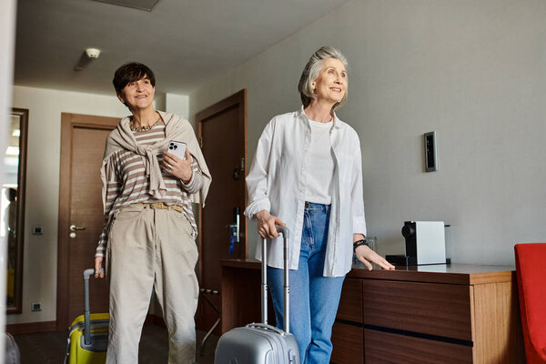 Senior lesbian couple stands with luggage in hotel room.