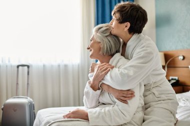 Elderly lesbian couple share a tender hug in a hotel room. clipart