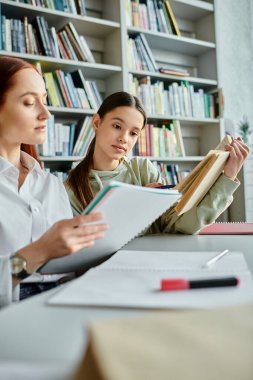 Kızıl saçlı bir öğretmen, okul sonrası kütüphane masasında ders veren genç bir kıza dizüstü bilgisayarla bilgi paylaşıyor..