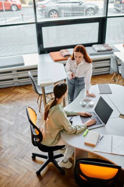 Kızıl saçlı öğretmen, genç kıza bir ofis ortamında ders veriyor. İkisi de modern eğitim için dizüstü bilgisayarda çalışmaya odaklanmış..