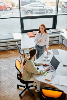 Kızıl saçlı bir öğretmen, genç bir kıza okul sonrası derslerinde modern ofis ortamında rehberlik ediyor..