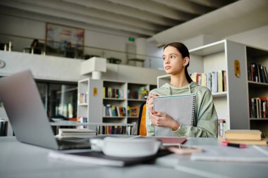 A teenage girl deep in thought as she works on her homework using a laptop in a quiet library setting. clipart