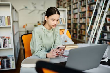 Genç bir kız, dizüstü bilgisayarıyla ev ödevlerine odaklanmış bir şekilde masada oturuyor ve cep telefonunda modern eğitimin güncellemelerini kontrol ediyor..