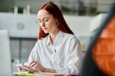 Kızıl saçlı bir bayan öğretmen çevrimiçi öğretmenlik yapıyor, bir masada oturuyor ve okuldan sonra akıllı bir telefonla iletişim kuruyor..