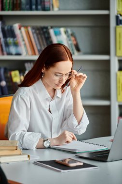 A red-haired female tutor is teaching online using a laptop in a library after a school lesson. clipart