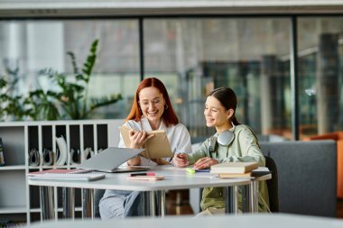 A redhead woman is tutoring a teenage girl at a table in an office, using a laptop for modern education after school lessons. clipart