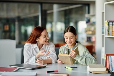 A redhead woman teaches a teenage girl in a library, engrossed in after-school lessons with a laptop. clipart