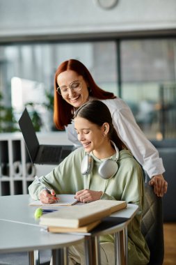 A redhead woman tutors a teenage girl at a table in an office, both focused on a laptop during after school lessons. clipart