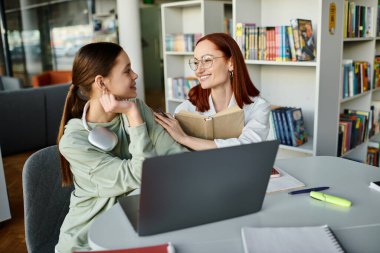 Bir öğretmen, kızıl saçlı bir kadın, okul sonrası ders sırasında genç bir kıza ders veriyor, ikisi de dizüstü bilgisayar kullanarak birlikte öğrenmekle meşgul..
