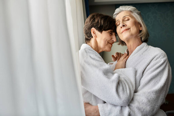 A senior lesbian couple, one in a bathrobe, hugging affectionately.