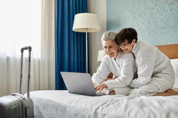 Senior lesbian couple share a laptop on a bed in a cozy moment.