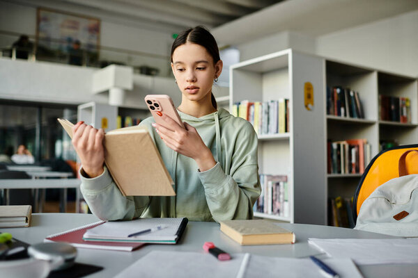 A teenage girl sits at a desk engrossed in a book while glancing at her phone, blending traditional study with modern distractions.