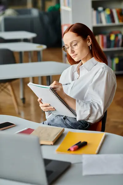 stock image A redheaded female tutor sits at her desk, focused on writing in a notebook after an online lesson.