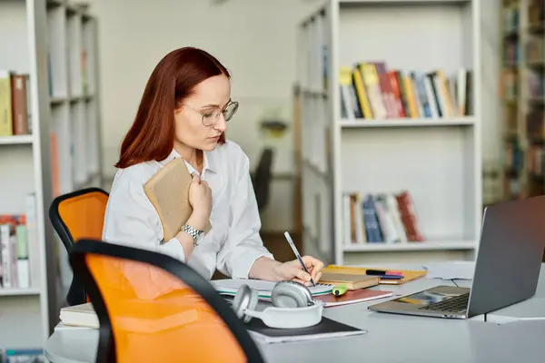 stock image A female tutor with red hair providing an after-school lesson online, using a laptop at a desk in a library setting.