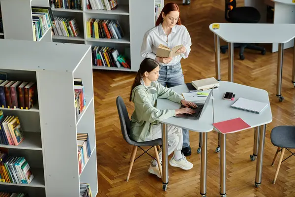 stock image A redheaded woman tutors a teenage girl, engaged in an after-school lesson using a laptop in a quiet library setting.